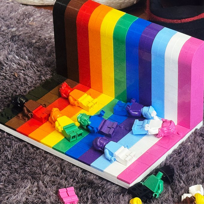 A vibrant rainbow-themed Lego display sits on a fuzzy gray rug, featuring small toy vehicles and figures arranged along colorful stripes, posted in honor of "VRM day." The Pride flag-inspired design celebrates visibility and inclusion within the VRM community.