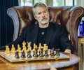 The image features an older man sitting in a chair with his legs crossed while playing chess. The man appears to be wearing a suit and tie, suggesting that he might have some professional background or formal attire. In front of him is a large chessboard, indicating that they are engaged in a game of chess at the moment.

The image also contains text that reads "$MSTR &