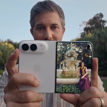 A man holds up a white foldable smartphone displaying an image of a woman in a sari posing in front of a tiered fountain.
