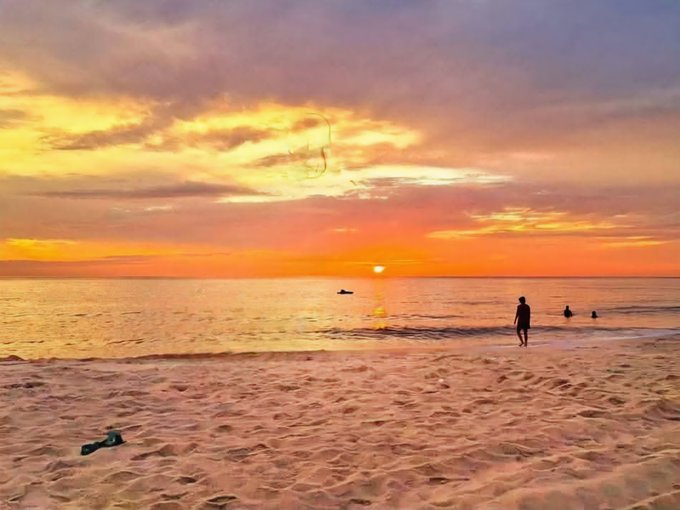 A serene beach sunset scene with orange and purple sky, a person silhouetted near the water, and swimmers visible in the ocean.