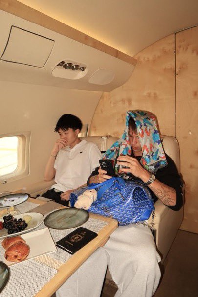 Two young men are seated in the luxurious cabin of a private jet, with one wearing a colorful patterned towel over his head while looking at a phone, surrounded by plates of food including fruit and pastries.