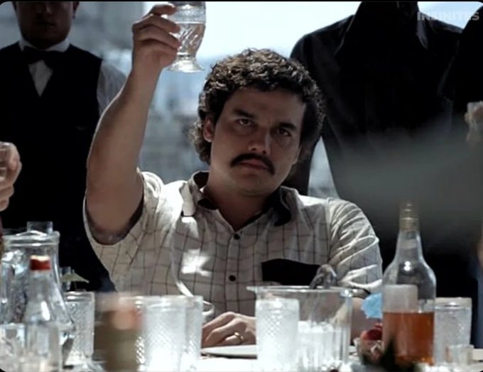 A scene from a film showing a mustached man in a checkered shirt raising a glass in a toast at a crowded table filled with bottles and glasses.