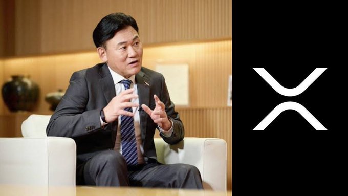 A man in a suit gestures while speaking during an interview, with the XRP logo prominently displayed on the right side of the image, visually connecting the executive's commentary to the cryptocurrency discussed in the post about Japanese banks adopting XRP within the $7 trillion banking system.