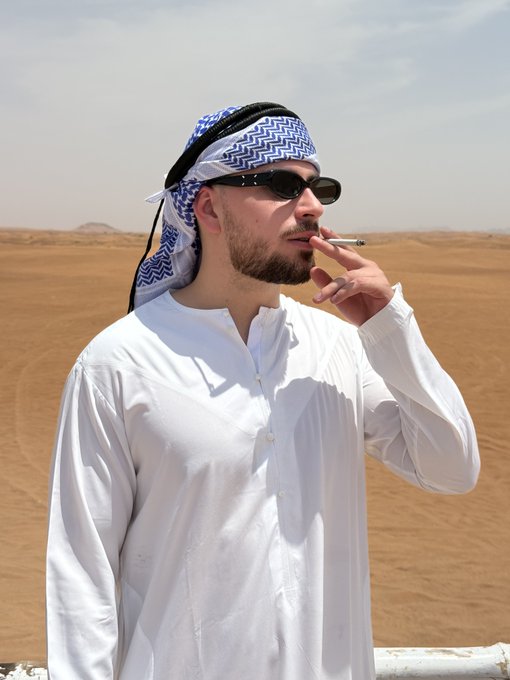 A man in traditional white Arab attire and a keffiyeh smoking a cigarette in a desert setting.