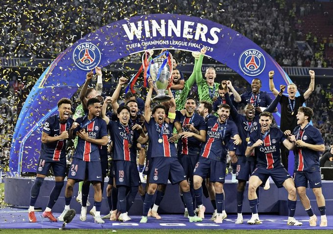 The image shows a group of soccer players celebrating their victory with high fives, hugs, and jumping up and down. They are wearing blue uniforms and holding a trophy aloft as they celebrate. The trophy has the words "Winners" written on it in large letters. In the background, there is an arch made of confetti that adds to the festive atmosphere.