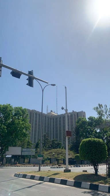 A street view on a sunny day showing a tall building, a traffic light, and trees under a clear blue sky.