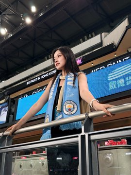 A young woman wearing a blue Manchester City football scarf standing behind a railing at what appears to be an indoor event venue.