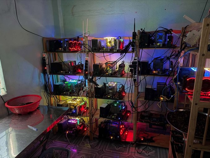 A dimly lit room contains multiple shelves densely packed with electronic equipment, tangled cables, and glowing components, suggesting a cryptocurrency mining setup.