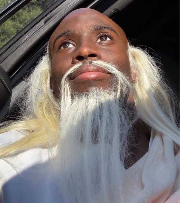 A person with a shaved head and a long white-blonde beard sits inside a vehicle, looking upward with a thoughtful expression.