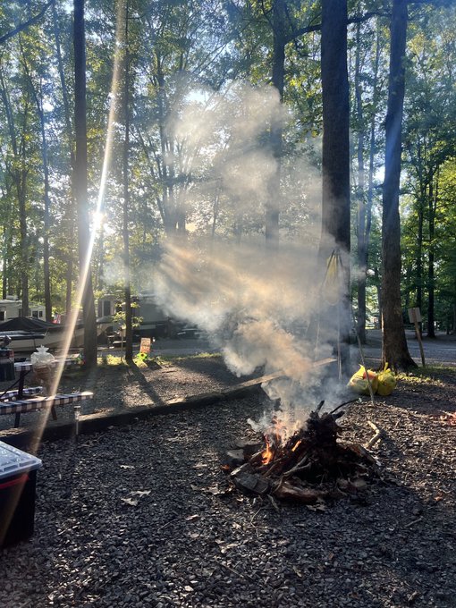 A campfire with smoke rising through sunlit rays at a wooded campground, with picnic tables, an RV, and yellow trash bags visible in the background.