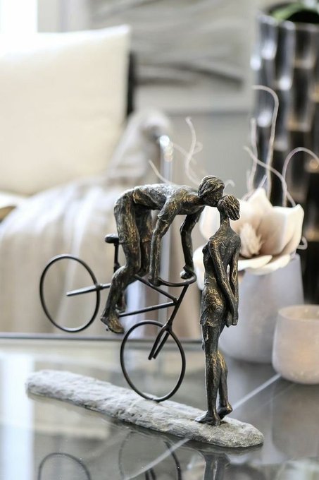 A decorative bronze sculpture on a glass table depicts two figures with a bicycle, one appearing to mount it while the other stands beside it, set in a softly lit room with pillows and a vase.