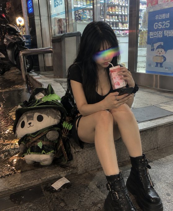 A young woman with long dark hair sits on a curb at night drinking from a juice box and looking at her phone, next to a panda plushie dressed as a hunter.