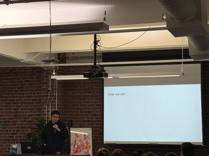 A man giving a presentation titled 'How we win' on a projector screen in a room with exposed brick walls and industrial ceiling fixtures.
