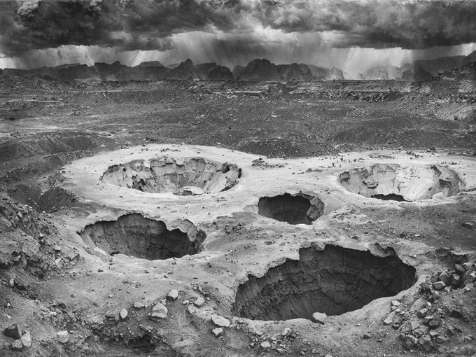 A black and white image of a barren landscape featuring several large craters or holes in the ground, with mountains and a dramatic cloudy sky in the background.