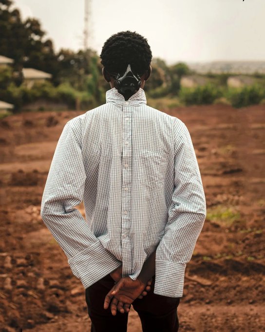 A person standing outdoors with their face covered by a black mask, wearing an oversized grey and white checkered shirt with their hands behind their back.