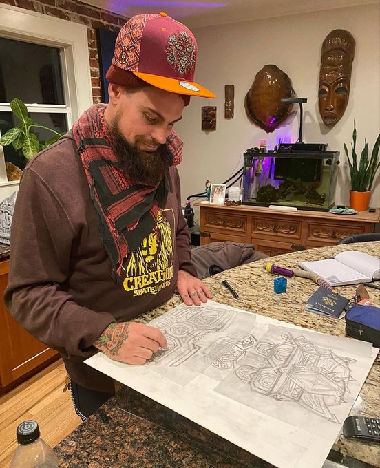 A bearded man wearing a red patterned cap and a brown 'Creature' sweatshirt works on a detailed pencil drawing of a geometric figure at a kitchen counter.
