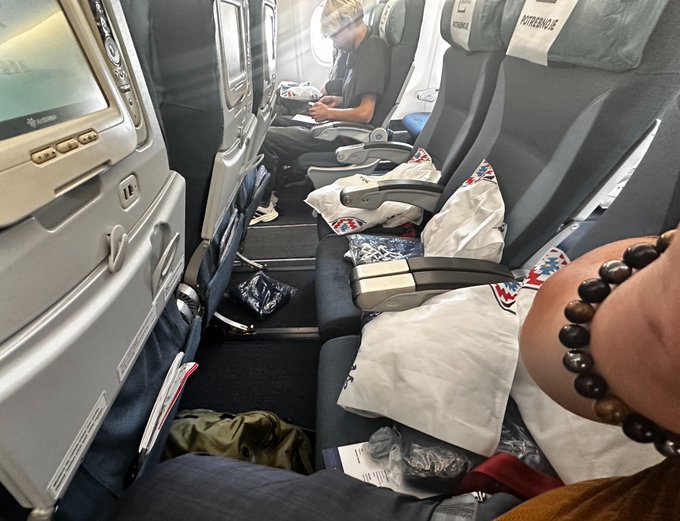 A messy airplane cabin interior with discarded food wrappers, pillows, and debris scattered across the seats, with a passenger wearing goggles looking at a device and a hand wearing a dark beaded bracelet visible in the foreground.