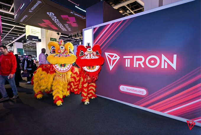 Two traditional Chinese lion dancers (one yellow, one red) pose in front of a large illuminated TRON (TRX) booth displaying the brand logo and "trondao.org" at a conference. The vibrant display reflects the post's message highlighting TRON's quiet but massive real-world activity—hundreds of millions of accounts and billions of transactions flowing through the network without needing hype.