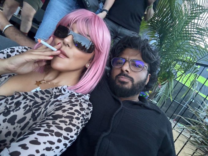 A selfie of a couple sitting together outdoors; the woman has bright pink hair, wears sunglasses, and is smoking a cigarette, while the man beside her has dark curly hair, glasses, and a beard.