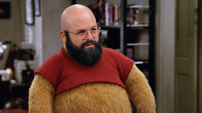 A bald man with a thick beard and glasses in a fur suit looks concerned in an office setting. The image ties to the meme about the ubiquity of memecoins, with his incredulous expression capturing the "Jerry" moment in a Seinfeld scene where he reacts to the overwhelming prevalence of crypto tokens.