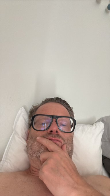A close-up selfie of a bearded man wearing black-framed glasses, lying against white pillows and resting his chin on his hand in a contemplative pose.