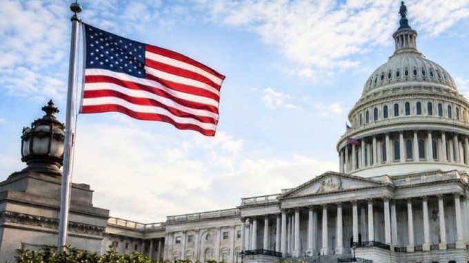 The image shows an American flag waving in front of the U.S. Capitol building's iconic white dome against a blue sky. It visually anchors the post's announcement about the departure of the White House's CLARITY Act negotiation lead, Patrick Witt, emphasizing the governmental and legislative backdrop of the Senate vote scheduled for July 20.