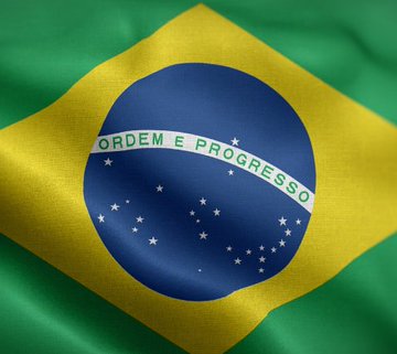 A waving Brazilian flag is shown, with its green field, yellow diamond, and blue globe clearly visible, including the national motto "ORDEM E PROGRESSO" and white stars. The image accompanies a post about Brazil's new cryptocurrency regulation imposing 24-hour delays on large transfers to self-custody or foreign wallets, visually representing the country at the center of the regulatory announcement.