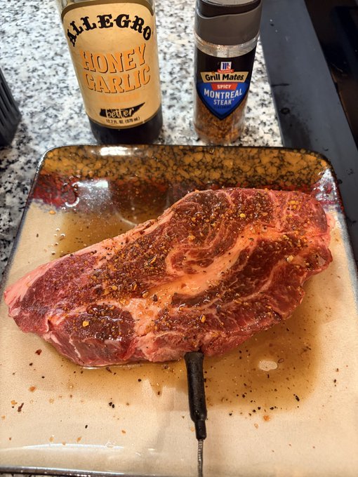A raw steak seasoned with spices sits on a square plate, with a meat thermometer inserted into it; behind the steak are two bottles of Allegro Honey Garlic marinade and McCormick Grill Mates Montreal Steak seasoning.