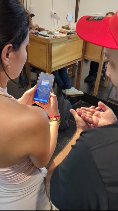 A woman showing her phone screen to a person wearing a red cap, seated in what appears to be a backstage or dressing room area.