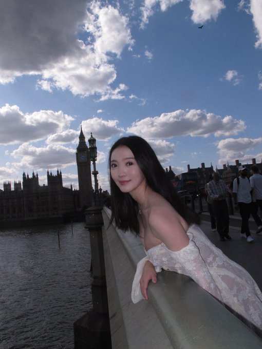 A woman in a white off-shoulder lace dress leans against a railing by the River Thames in London, with Big Ben and the Houses of Parliament visible behind her under a partly cloudy blue sky.