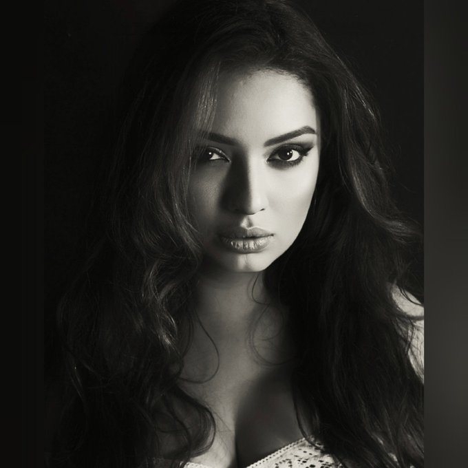 A black and white portrait of a young woman with long dark wavy hair, heavy eye makeup, and a white top against a dark background.