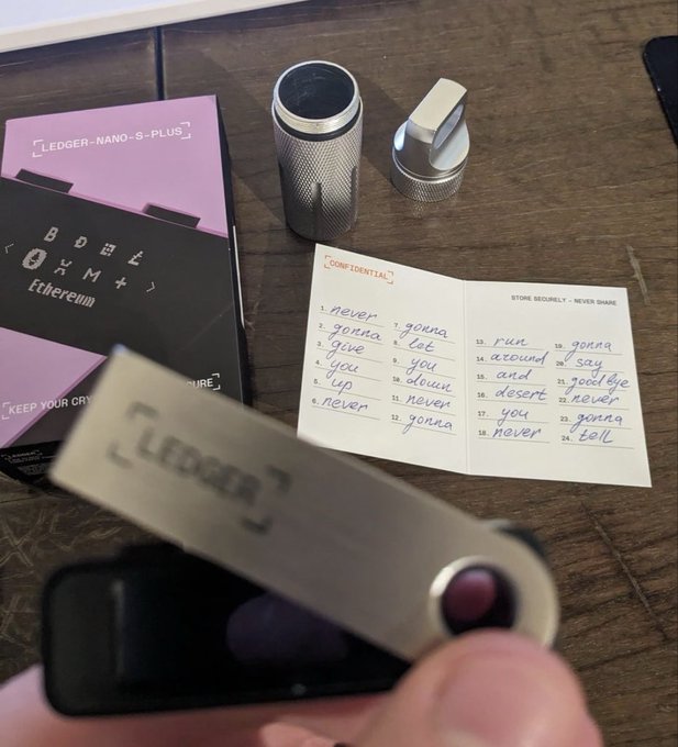 A hand holds a silver Ledger hardware wallet in the foreground, with its packaging ('LEDGER NANO S PLUS', 'BODEE', 'Ethereum') and a 'STONE SECRETLY - NEVER SHARE' recovery seed booklet visible on a table in the background.
