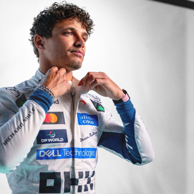 A young man with curly hair adjusts his racing suit, which features sponsor logos including "Cisco," "DP World," "Dell Technologies," "Richard Mille," and the number "4," while looking off into the distance—capturing the focused intensity of a Formula 1 driver preparing for the European racing circuit.