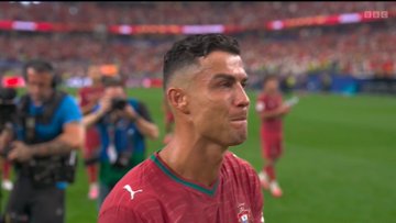 A close-up of soccer player Cristiano Ronaldo in a Portugal jersey on the field with photographers and a stadium crowd in the background.