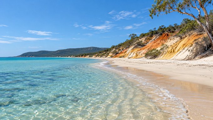 A pristine beach with turquoise water gently lapping against white sand, bordered by colorful red and orange sandstone cliffs under a bright blue sky.