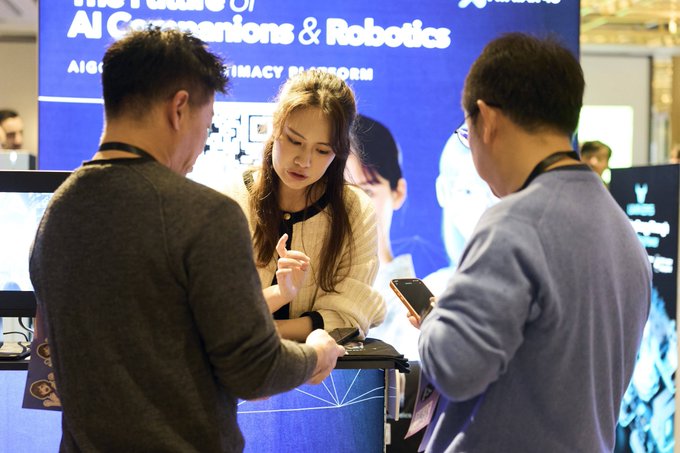 A woman with long hair stands between two men at a tech exhibition booth, with a banner behind them partially reading 'The Future of AI Companions & Robotics'.