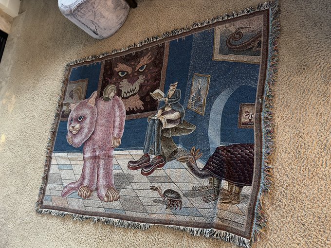 A woven fringed tapestry blanket featuring whimsical surreal artwork of a pink bear-like figure, a beast portrait, a seated figure in a hat, and a large bird, laid out on a carpeted floor. The image likely showcases one of the giclée prints given as a reward in the Beaks raffle, matching the surreal illustrative style of the prizes mentioned.