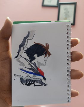 The image features a hand holding an open spiral-bound notebook with a drawing of a person's face inside. The notebook has a green cover and white pages, giving it a unique appearance. The drawing depicts a person wearing a red shirt and blue pants against a pink background. The caption below the image reads "We're Ready The official mint link has been shared, and the countdown has begun. Mint Date: June 5 Supply:  700 Leaders Mint Fee: 0.02 ETH Chain: Ethereum".