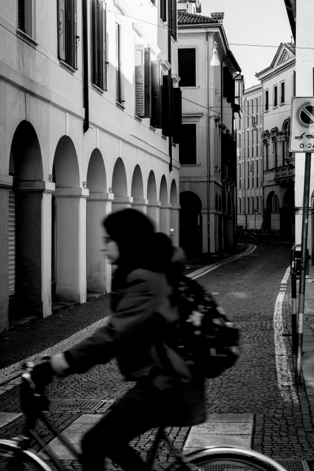 A black and white photograph showing a person riding a bicycle through a narrow European street lined with arched buildings.