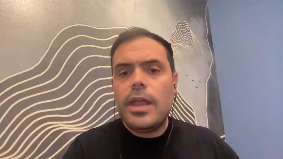 The image shows a man wearing headphones with his face partially visible as he speaks or looks at something off camera. The background features a blue wall with white lines, which appears to be an abstract art piece. The caption of the image reads "focus on what is in the image and how it relates to the post-quantum era and rn, you can't do this on any other L1".