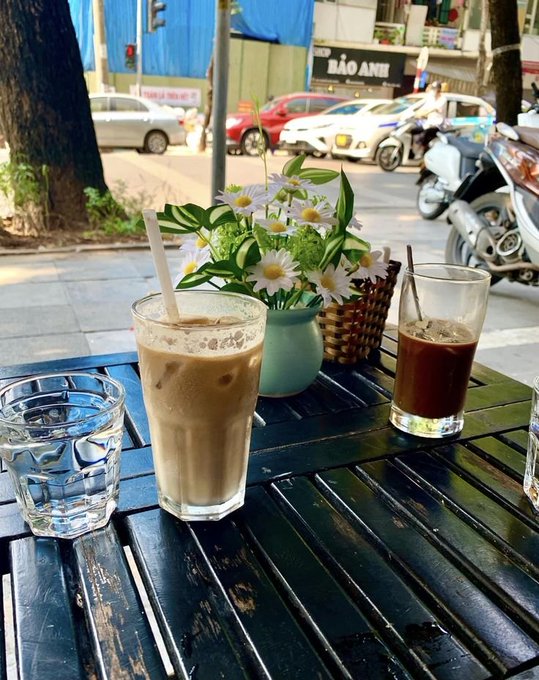 Two iced drinks and a glass of water sit on a black wooden table next to a small vase of white daisies on a city sidewalk with cars and scooters passing by.