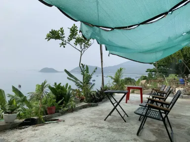 A serene outdoor patio with a green shade canopy, folding tables and chairs overlooking a calm bay with small islands, framed by potted plants.