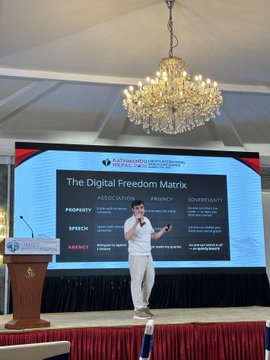 A man in white attire speaks on stage at a Kommando Strategic Conference, presenting a slide titled 'The Digital Freedom Matrix' that categorizes associations between Prosperty, Speech, Agency, Privacy, and Sovereignty under headings like 'ASSOCIATION' and 'GOVERNANCE.'