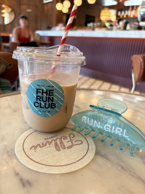 The image shows a table with two items on top of it - a plastic cup filled with coffee or tea, and a blue hair accessory (possibly a hair clip) placed next to it. The background reveals that this scene takes place at a restaurant or cafe setting. The caption "Sunday encrypted run with @fhenix x MYLE done" suggests that the person who set up the table might be participating in an online challenge or event, possibly related to running or fitness.