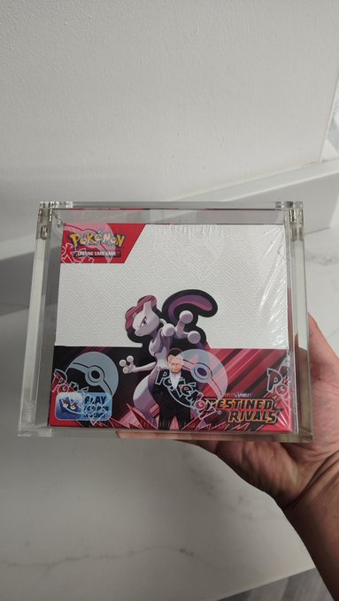 A hand holds a Pokémon Trading Card Game binder in clear plastic packaging, displaying artwork of a female character with 'PLAY! POKÉMON' and 'TRAINED RIVALS' branding visible.
