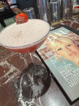 A pink cocktail garnished with a strawberry sits on a marble bar top next to an issue of T: The New York Times Style Magazine featuring Drew Barrymore.