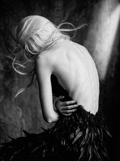 A black-and-white artistic photograph showing the back of a blonde woman with bare shoulders and a dark feathered garment, her head turned away.