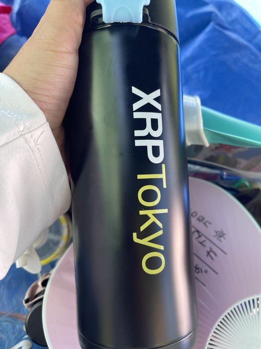 A hand holds a black water bottle with a blue cap, prominently displaying the text 'XRP TOKYO' in white and yellow letters.