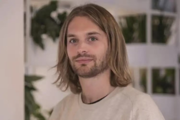 The image features a man with long hair wearing a white shirt who appears to be smiling at the camera. The background of the photo shows a wall adorned with various plants, giving off an impression of a well-decorated room or office space. There are several potted plants in different sizes and shapes scattered throughout the scene, adding a touch of greenery to the environment. The man's position in front of these plants suggests that he might be in a workspace or a shared living area where people can enjoy the natural ambiance created by the plants.