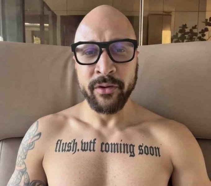 A bald man with glasses and a beard has 'flush.lol coming soon' tattooed across his chest, with another tattoo visible on his arm.